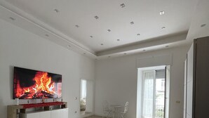 Amplia sala de estar con un televisor de pantalla plana grande que muestra una chimenea, un conjunto de sofás, un área de comedor con sillas y un gran ventanal corredizo que da a un balcón exterior.