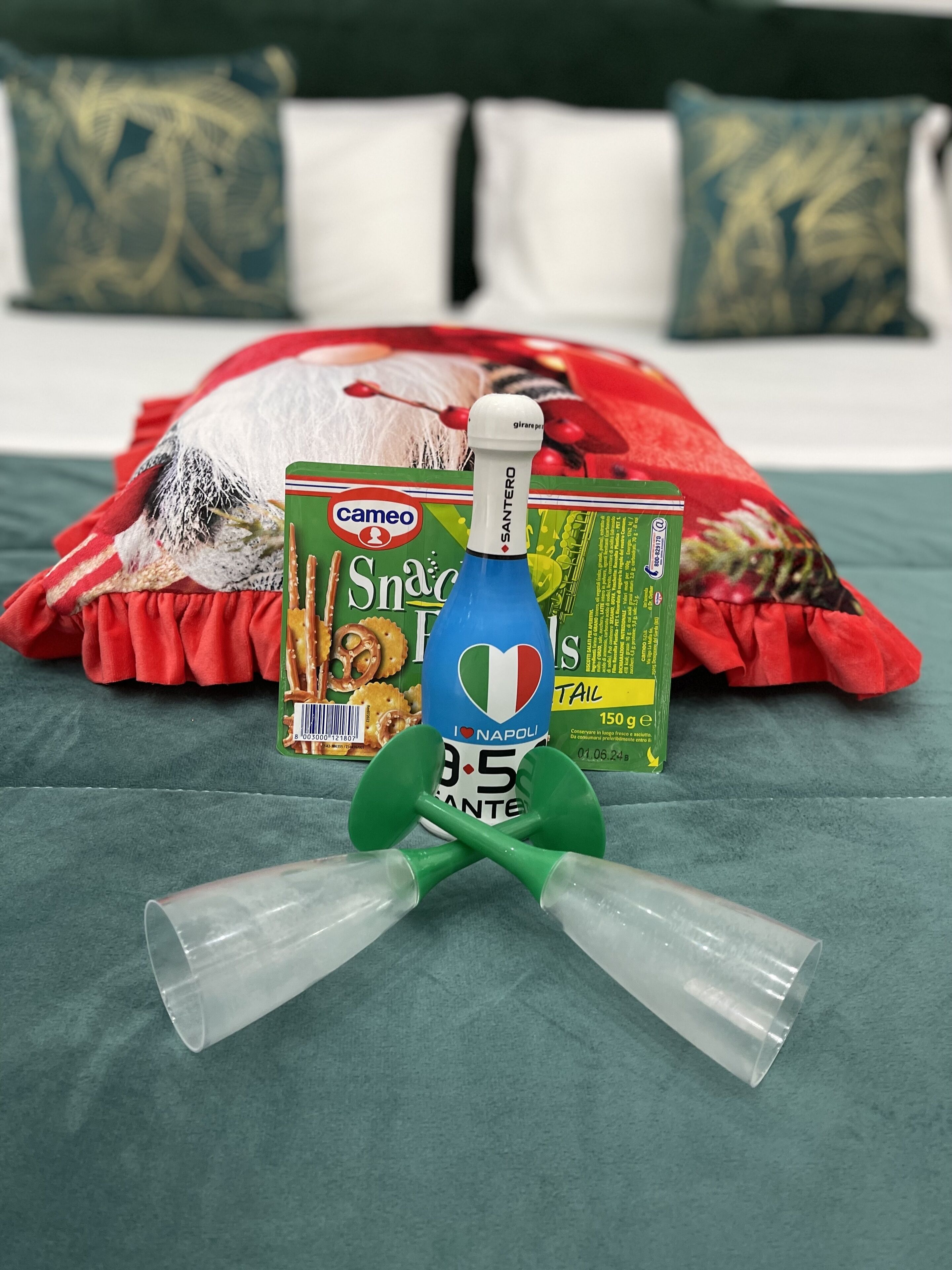 A bed with a red blanket and pillows, a green package labeled "Cameo," a bottle with "I love Napoli" and "3.5" on it, and two green plastic cups.