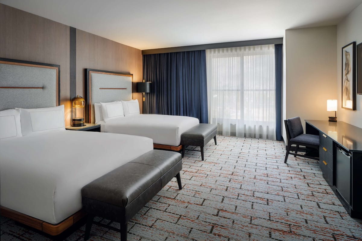 A hotel room with two beds, a desk, a chair, and a large window with curtains.