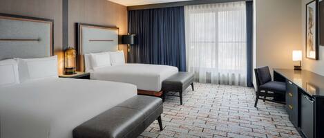 Habitación de hotel con dos camas, un escritorio, una silla y un ventanal con cortinas.
