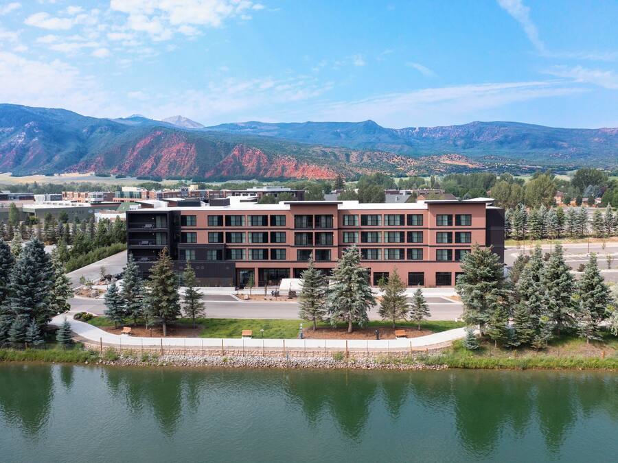 Un edificio moderno con techo plano, rodeado por un lago y árboles, con montañas al fondo.