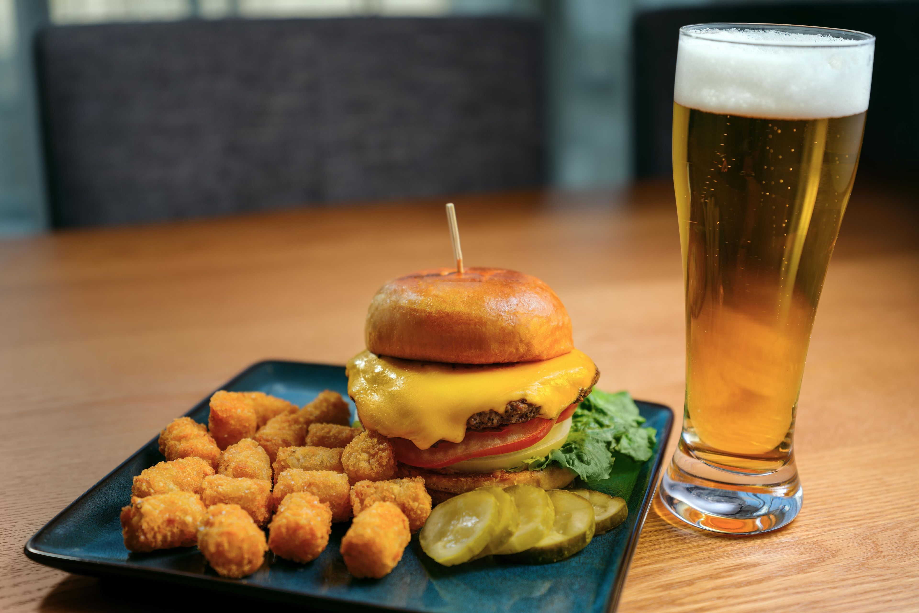A meal consisting of a burger, tater tots, and a pickle, accompanied by a glass of beer on a wooden table.