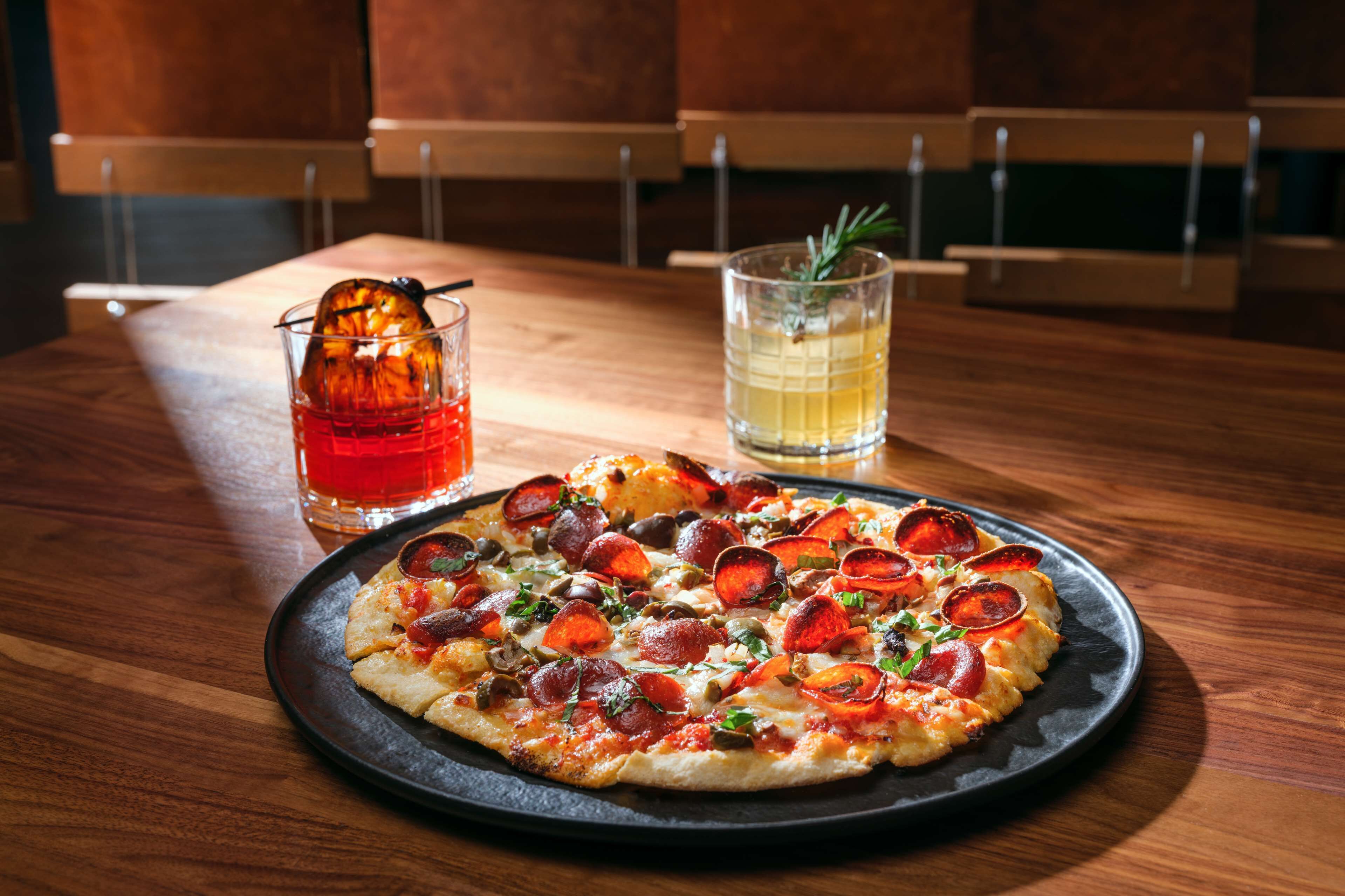 A pizza with pepperoni and mushrooms on a black plate, a glass of red drink, and a glass of yellow drink on a wooden table.