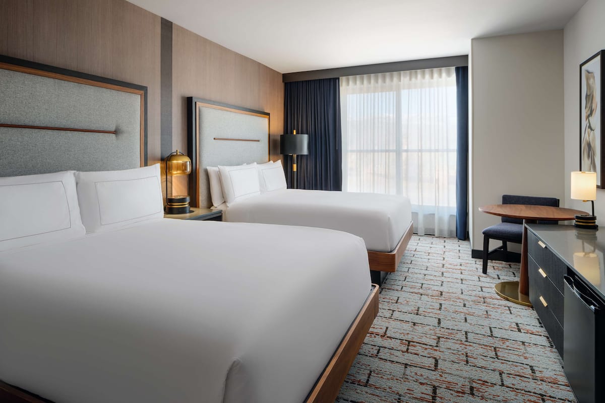 A hotel room with two beds, a desk, a chair, a lamp, and a window with curtains.