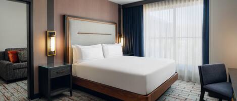 A hotel room with a large bed, a bedside table, a chair, a desk, a mirror, and a window with curtains.