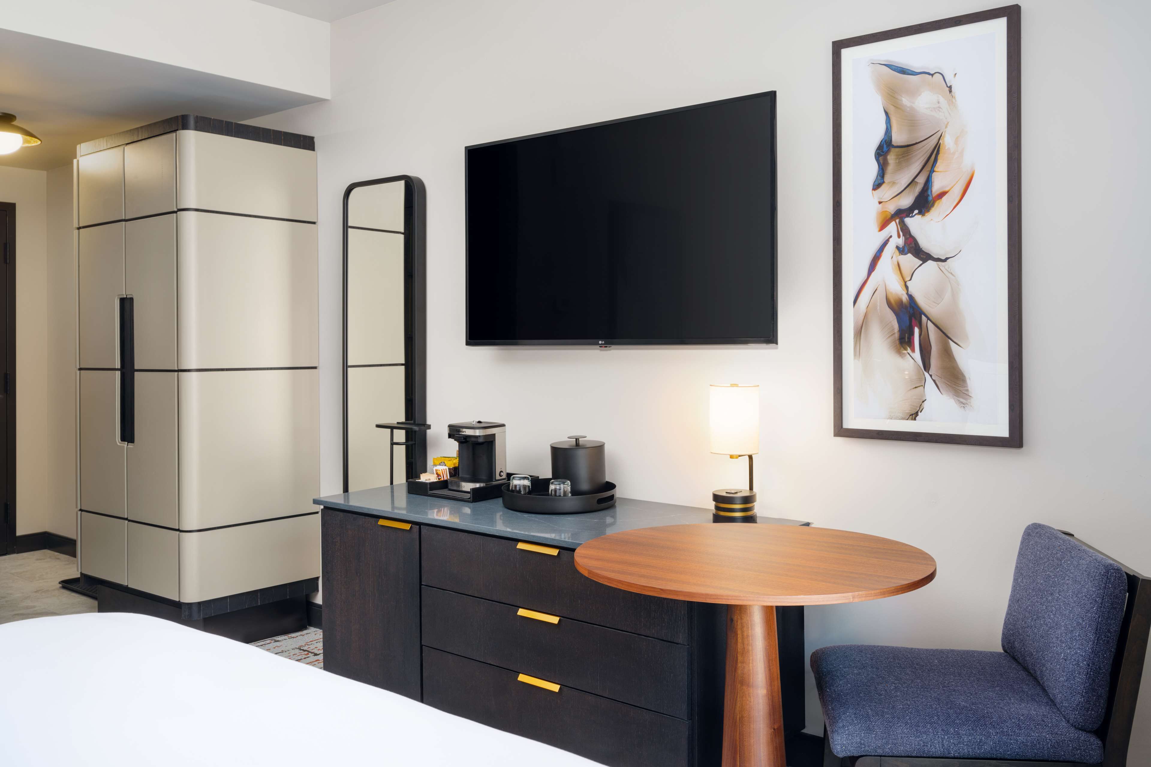 Habitación de hotel con una cama, un escritorio con cafetera, un televisor de pantalla plana, un cuadro abstracto enmarcado y una silla.