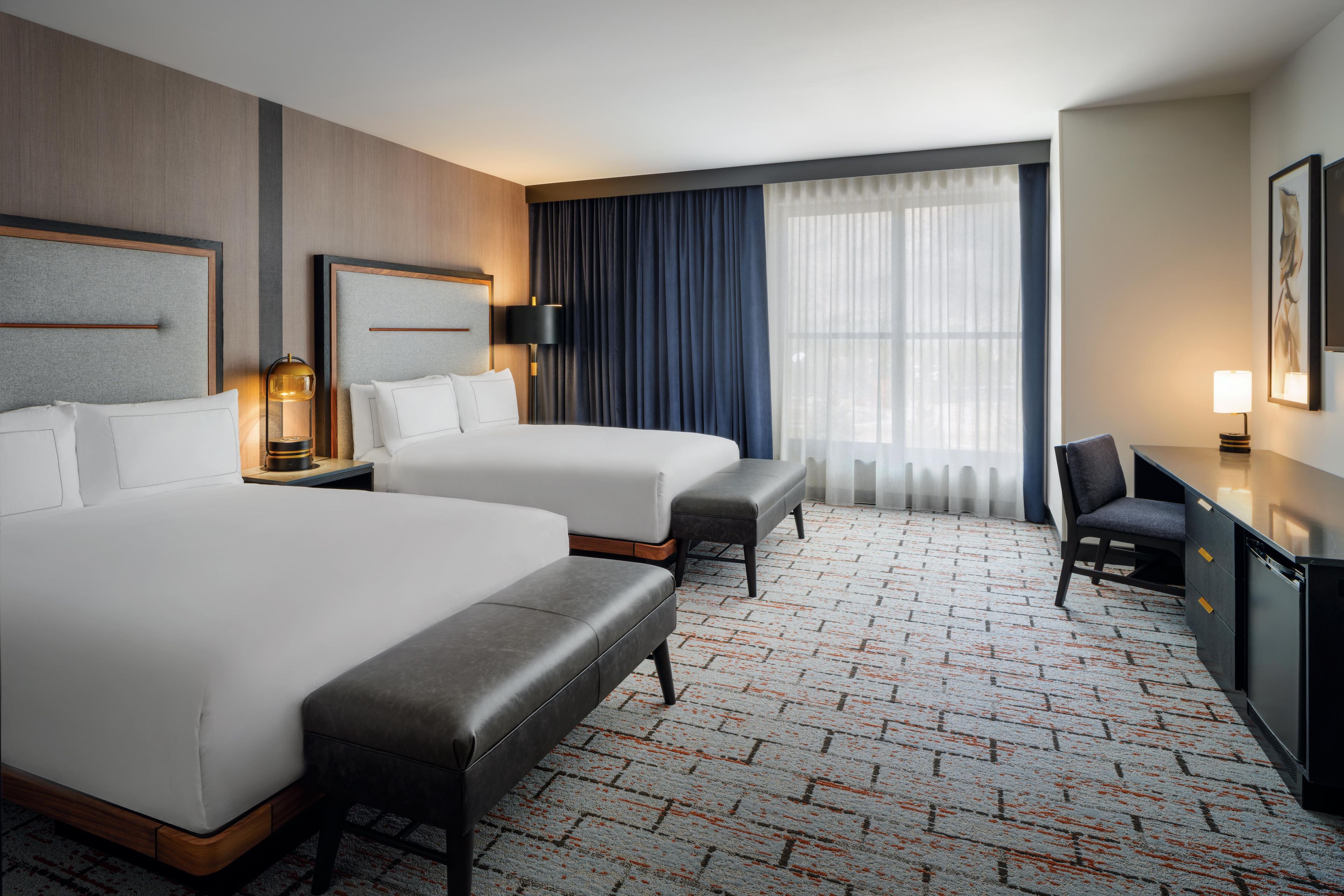 Ein Hotelzimmer mit zwei Betten, einem Schreibtisch, einem Stuhl und einem großen Fenster mit Vorhängen.