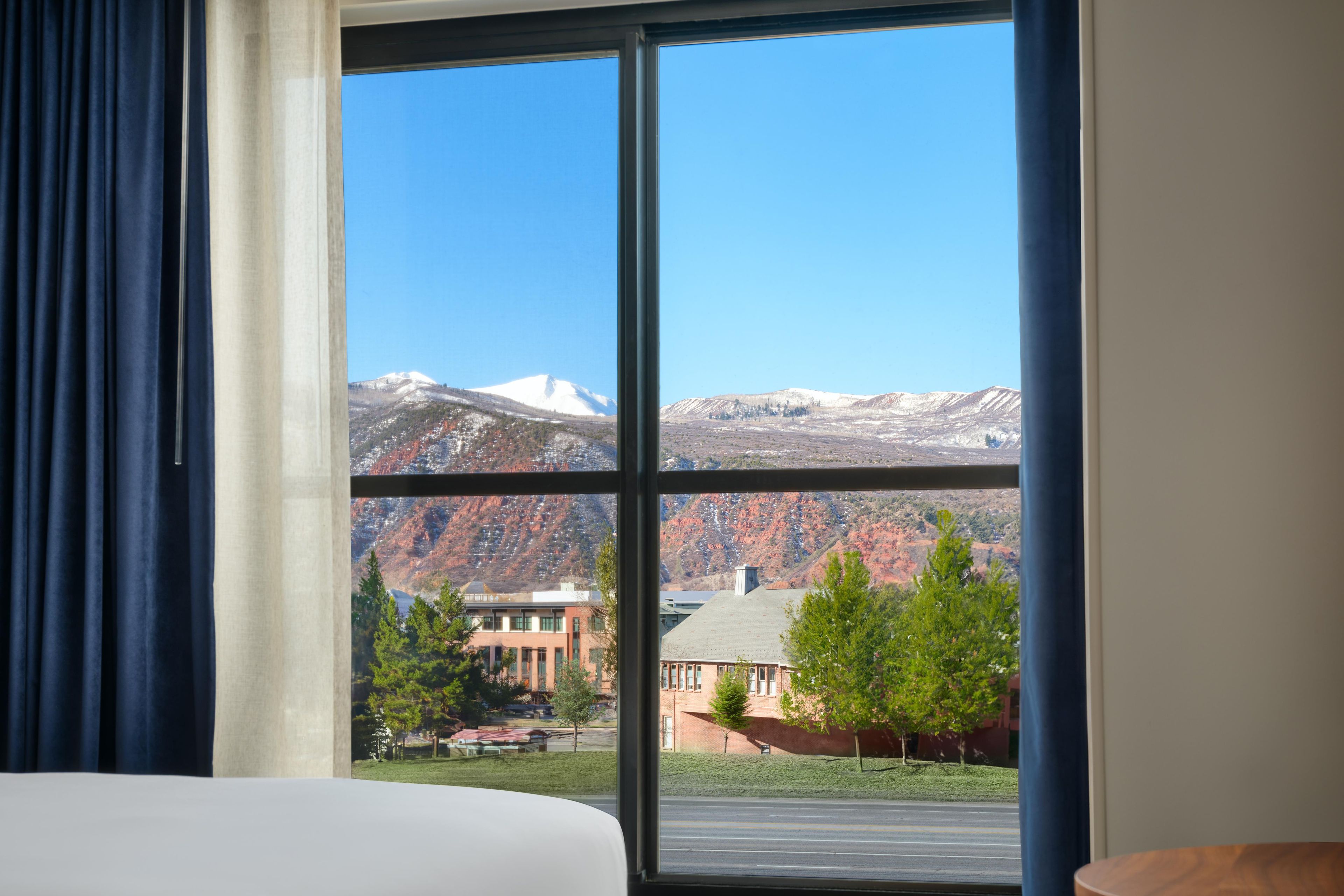 Habitación de hotel con un ventanal que ofrece vistas a una cadena montañosa y un edificio con techo rojo.