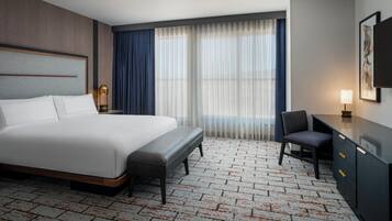 Habitación de hotel con una cama grande, un escritorio, una silla, una mesita de noche y un ventanal con cortinas.