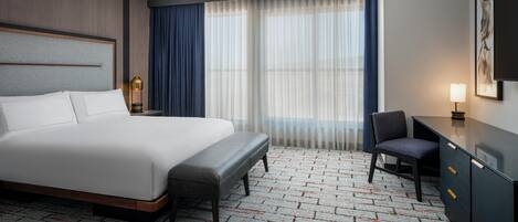 A hotel room with a large bed, a desk, a chair, a nightstand, and a window with curtains.