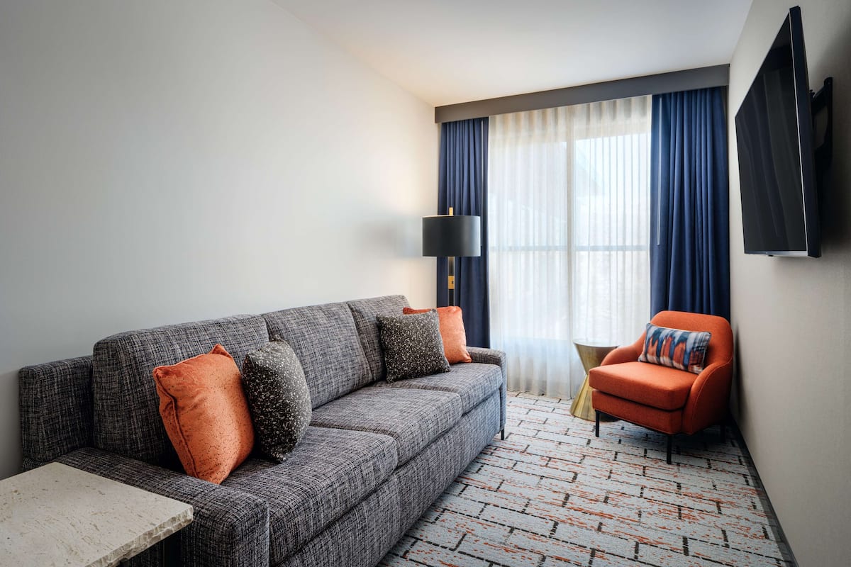 A modern living room with a grey sofa, orange cushions, a red armchair, a flat-screen TV, and a patterned rug.