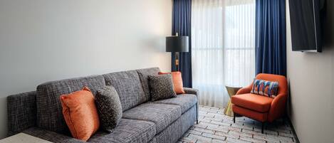Una sala moderna con un sofá gris, cojines naranja, un sillón rojo, un televisor de pantalla plana y una alfombra estampada.