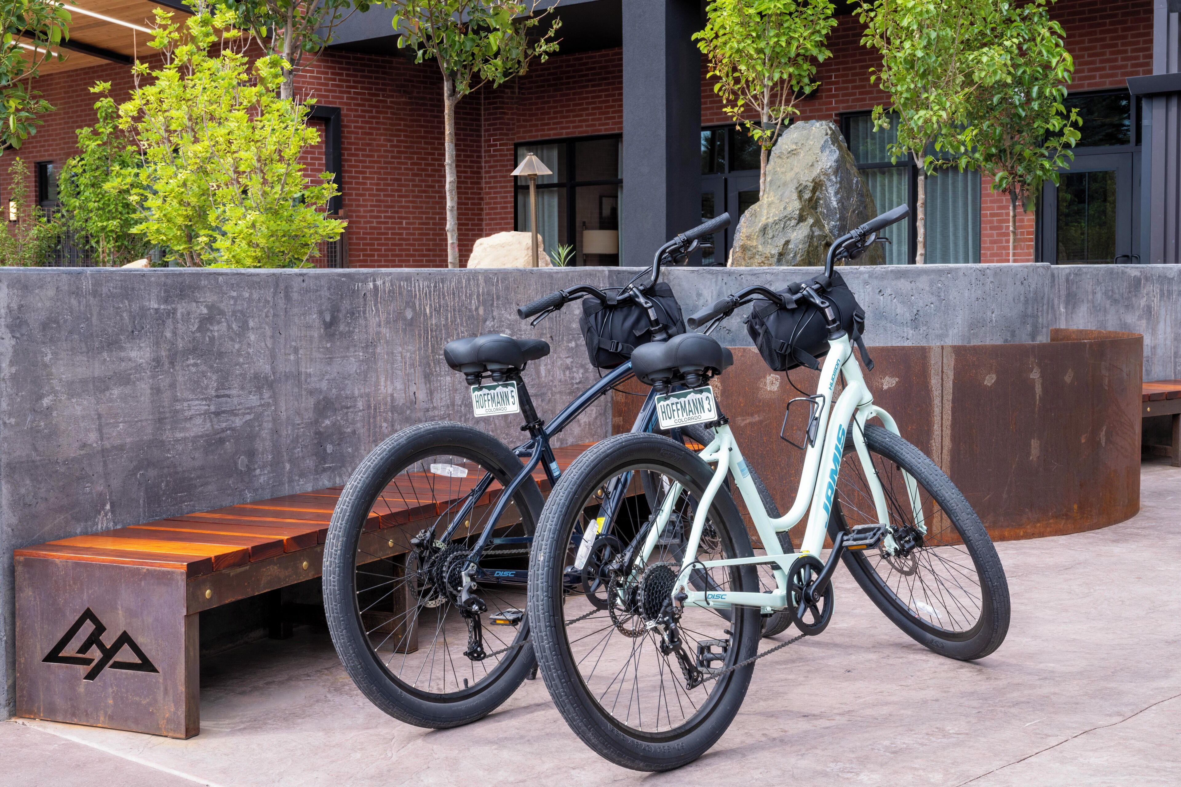 Deux vélos sont garés à côté d’un banc dans un espace extérieur, avec un bâtiment en briques et des arbres en arrière-plan.