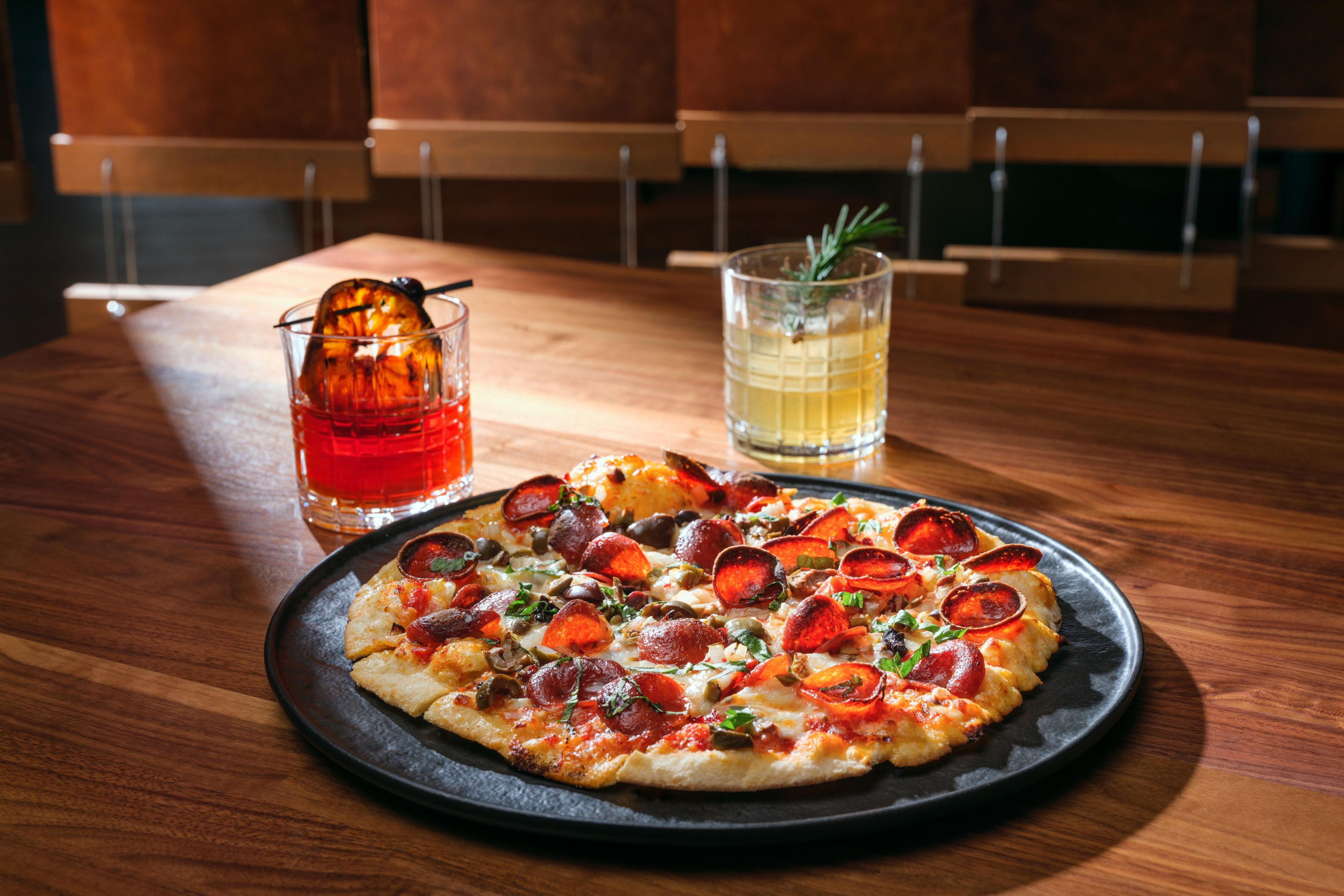 Eine Pizza mit Peperoni und Gemüse auf einem schwarzen Teller auf einem Holztisch.