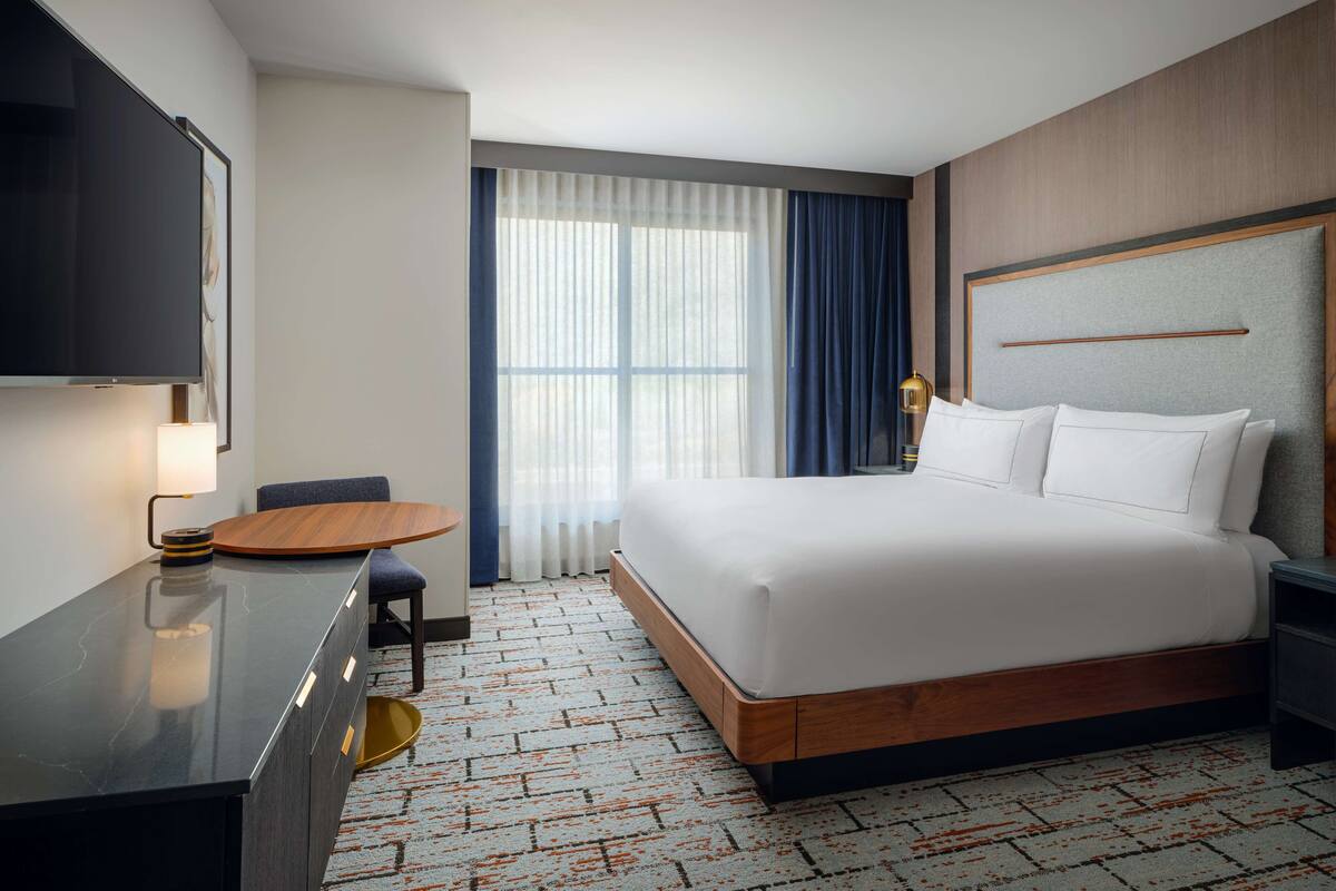 A hotel room with a large bed, a desk, a chair, a TV, and a window with curtains.