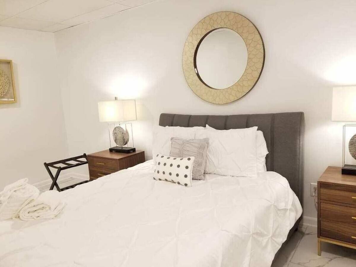 A neatly made bed with white bedding, a round mirror on the wall, and two bedside tables with lamps.