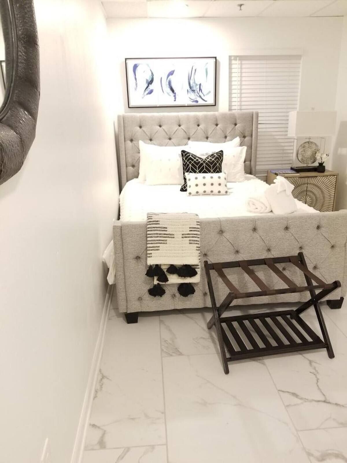 Un dormitorio con cabecera capitonada, una cama con ropa de cama blanca, una mesita de noche con lámpara y una obra de arte enmarcada sobre la cama.