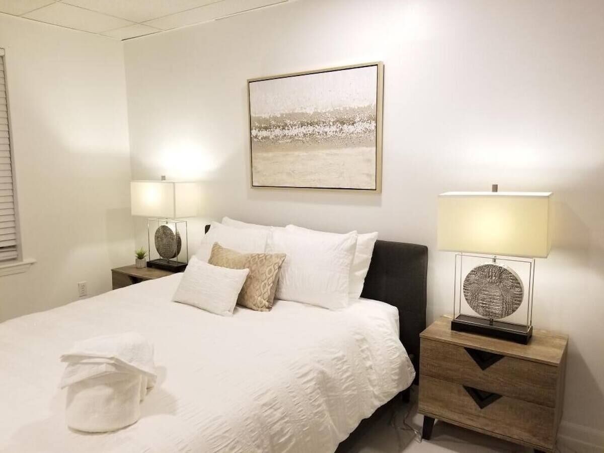 A neatly made bed with white linens and a folded towel, two bedside tables with lamps, a wall-mounted artwork, and a window with blinds.
