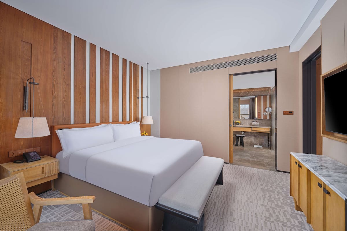 A modern hotel room with a large bed, bedside tables, a desk, a chair, a bench, and a TV.