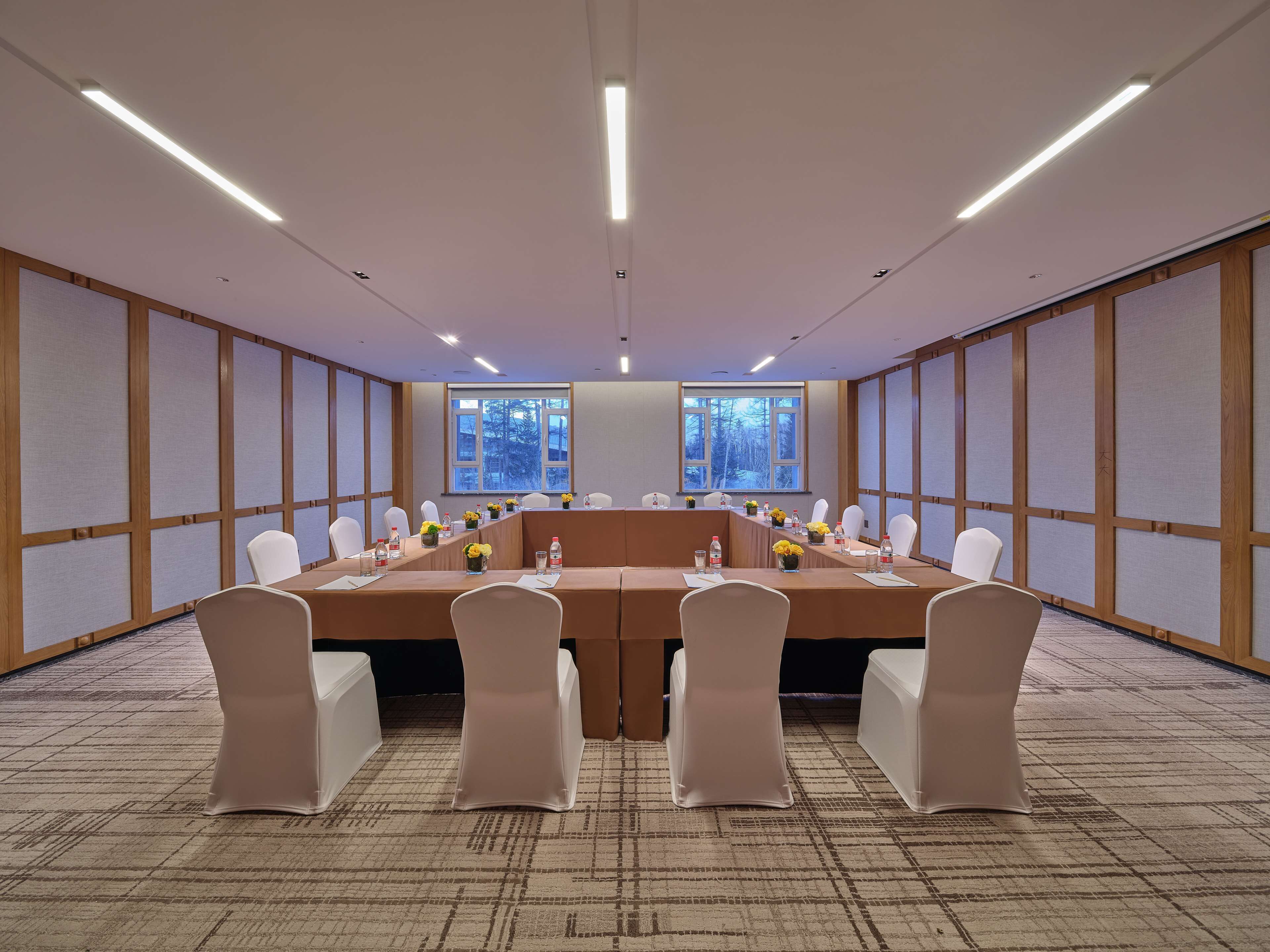 Une salle de conférence avec une longue table, des chaises blanches et une vue sur les arbres à travers de grandes fenêtres.