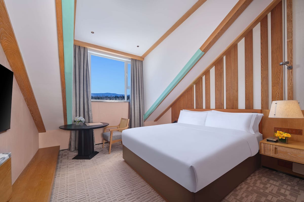 A hotel room with a large bed, a small table with a chair, a TV, and a window with curtains.
