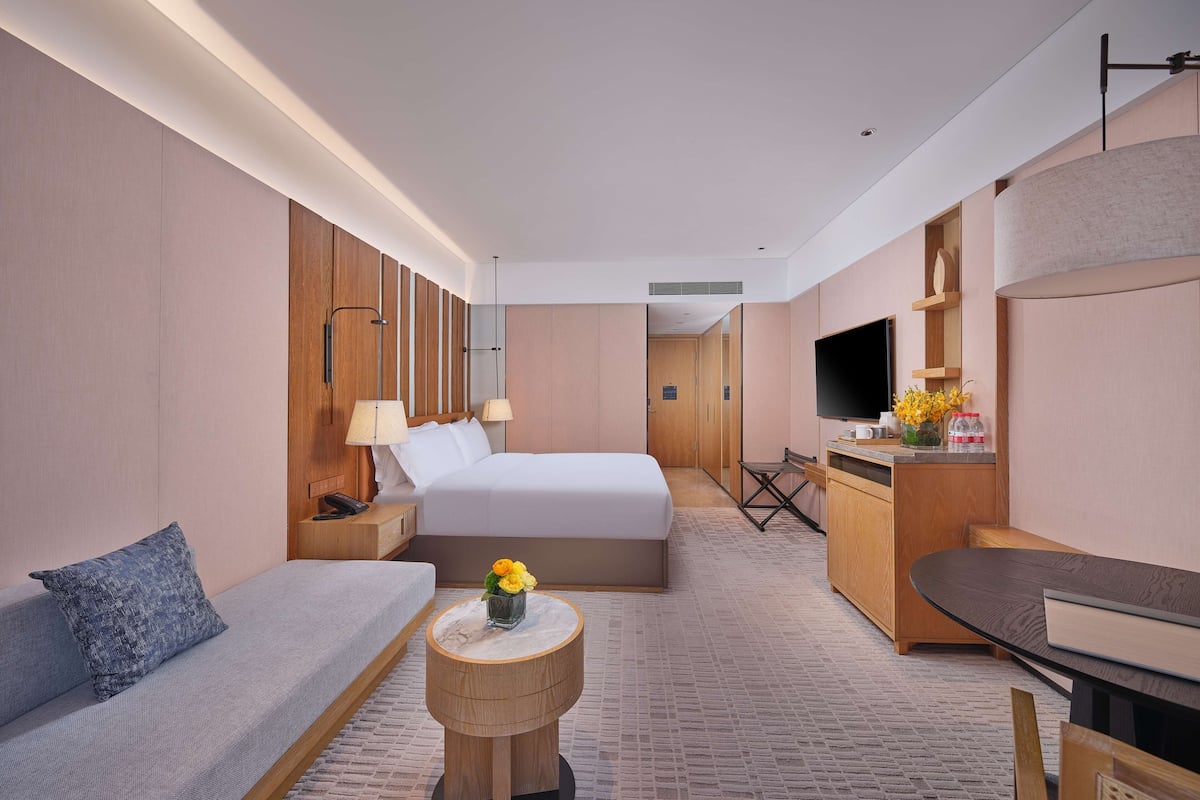 A hotel room with a bed, a sofa, a small round table with a vase, a desk with a chair, and a television.