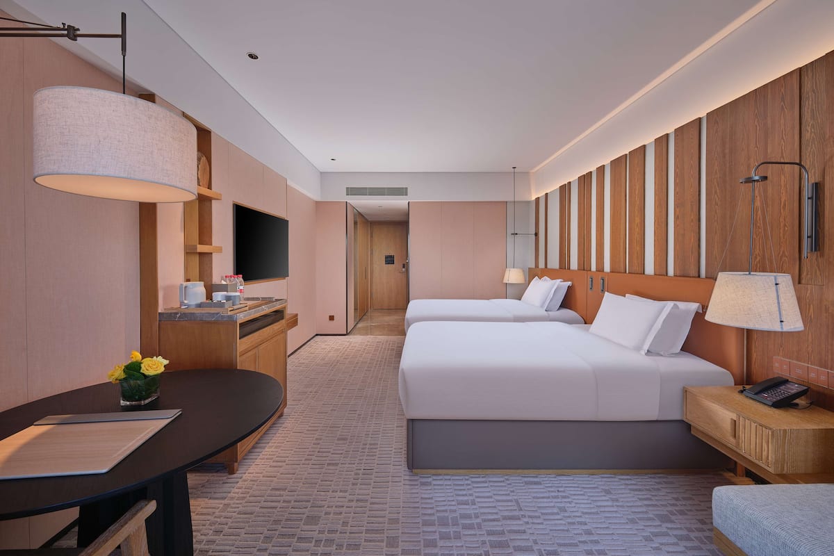 A hotel room with two beds, a desk, a chair, a TV, and a bathroom with a shower.