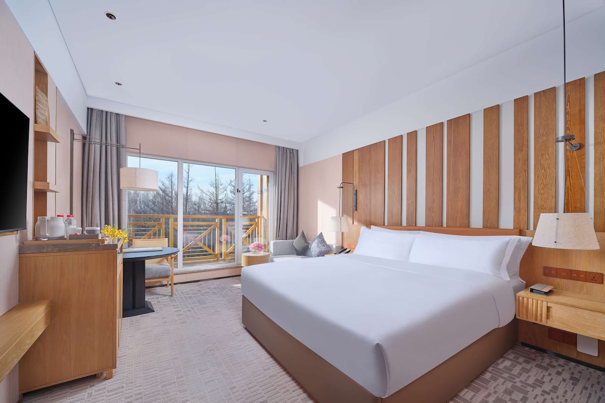 A modern hotel room with a large bed, a desk, a chair, a TV, and a balcony with a view.