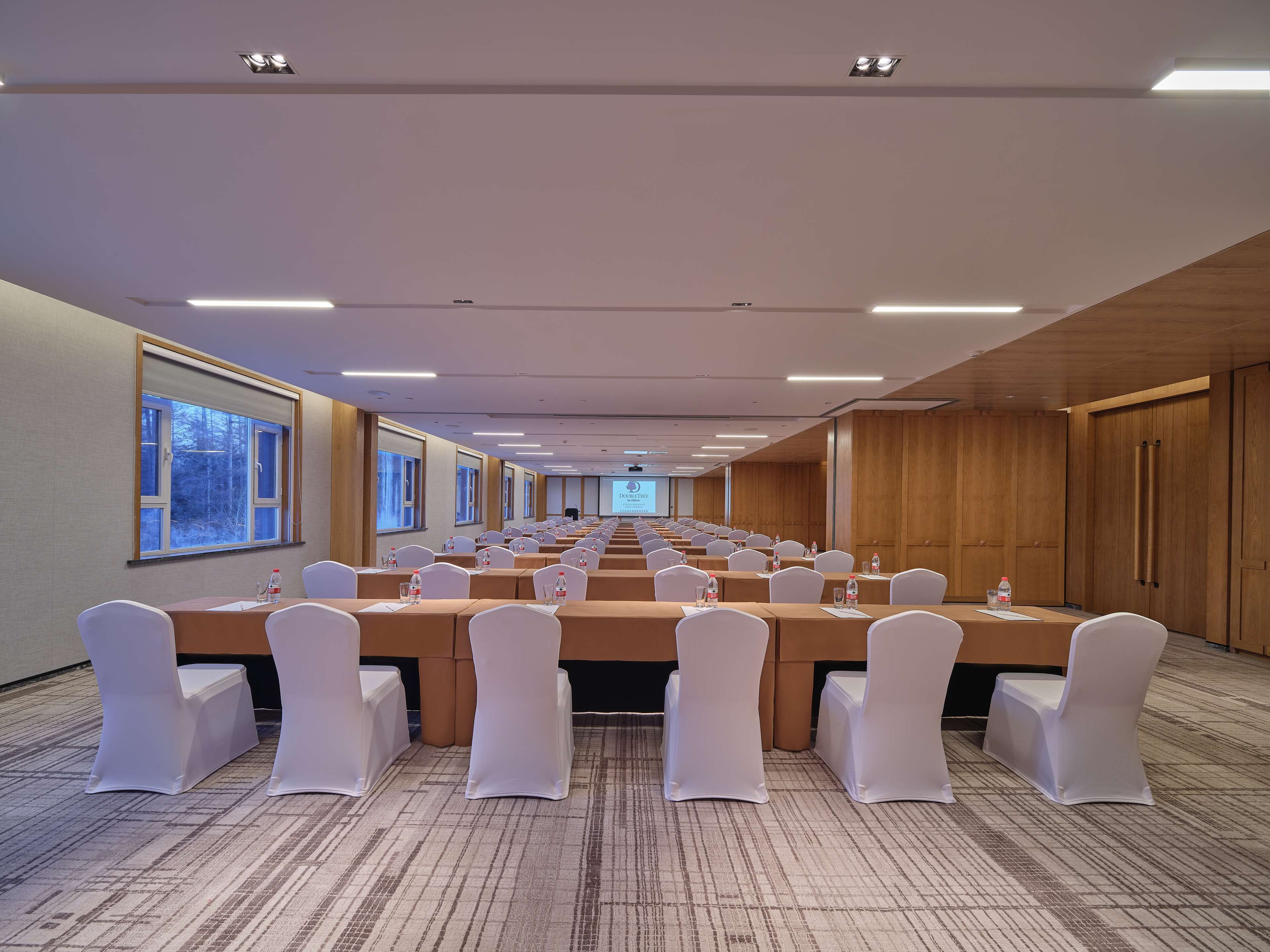 A conference room with rows of tables and chairs, a projector screen, and a large window with a view of trees.