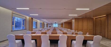 A conference room with rows of tables and chairs, a projector screen, and a large window with a view of trees.