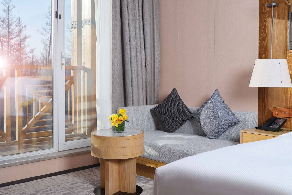 A hotel room with a bed, a sofa, a small wooden side table with a vase of flowers, a lamp, and a view of a wooden staircase outside.