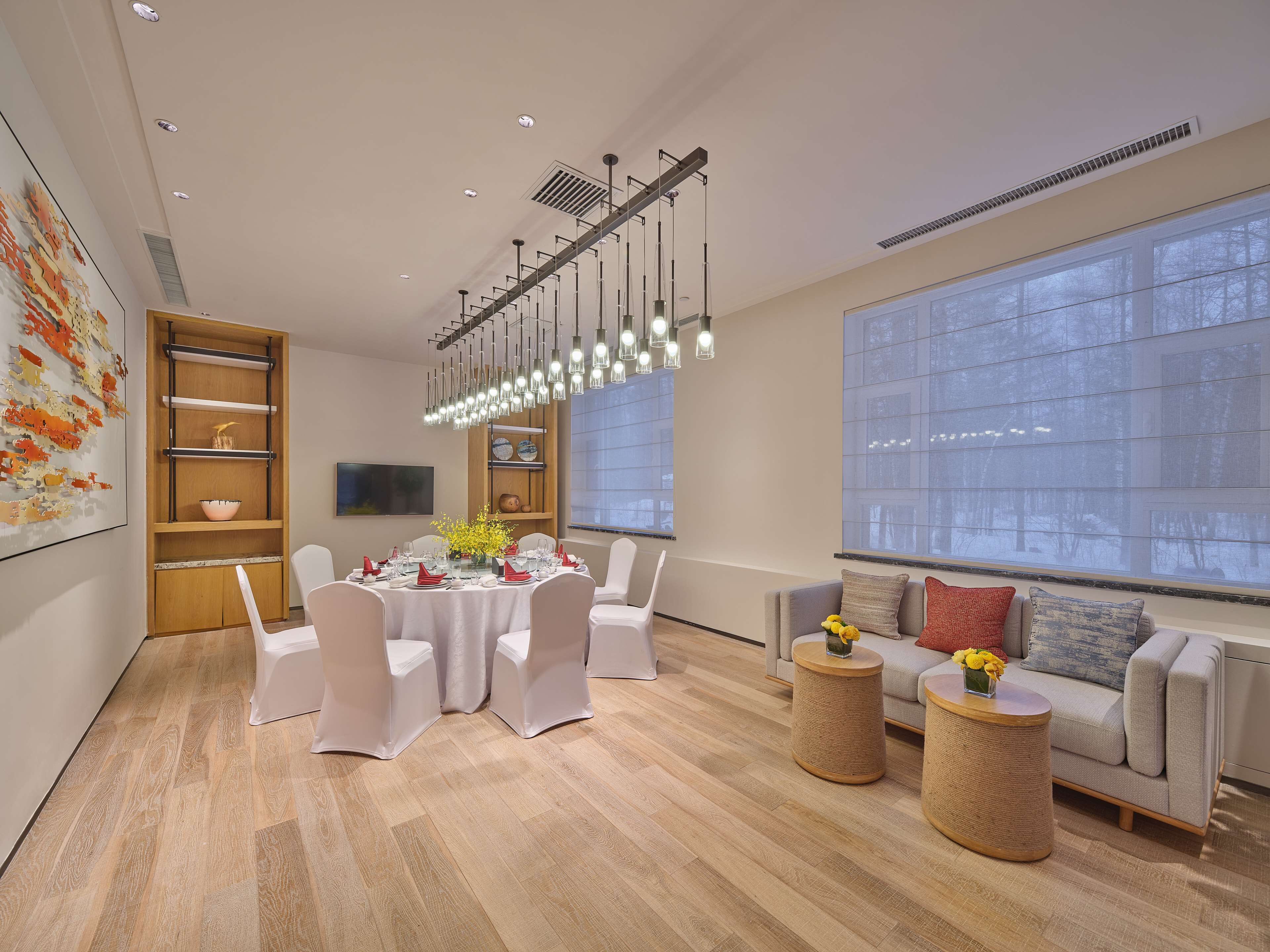 A modern dining area with a round table set for six, surrounded by white chairs. There is a wooden bookshelf with decorative items, a large window with a view of trees, and a grey sofa with a cushion.