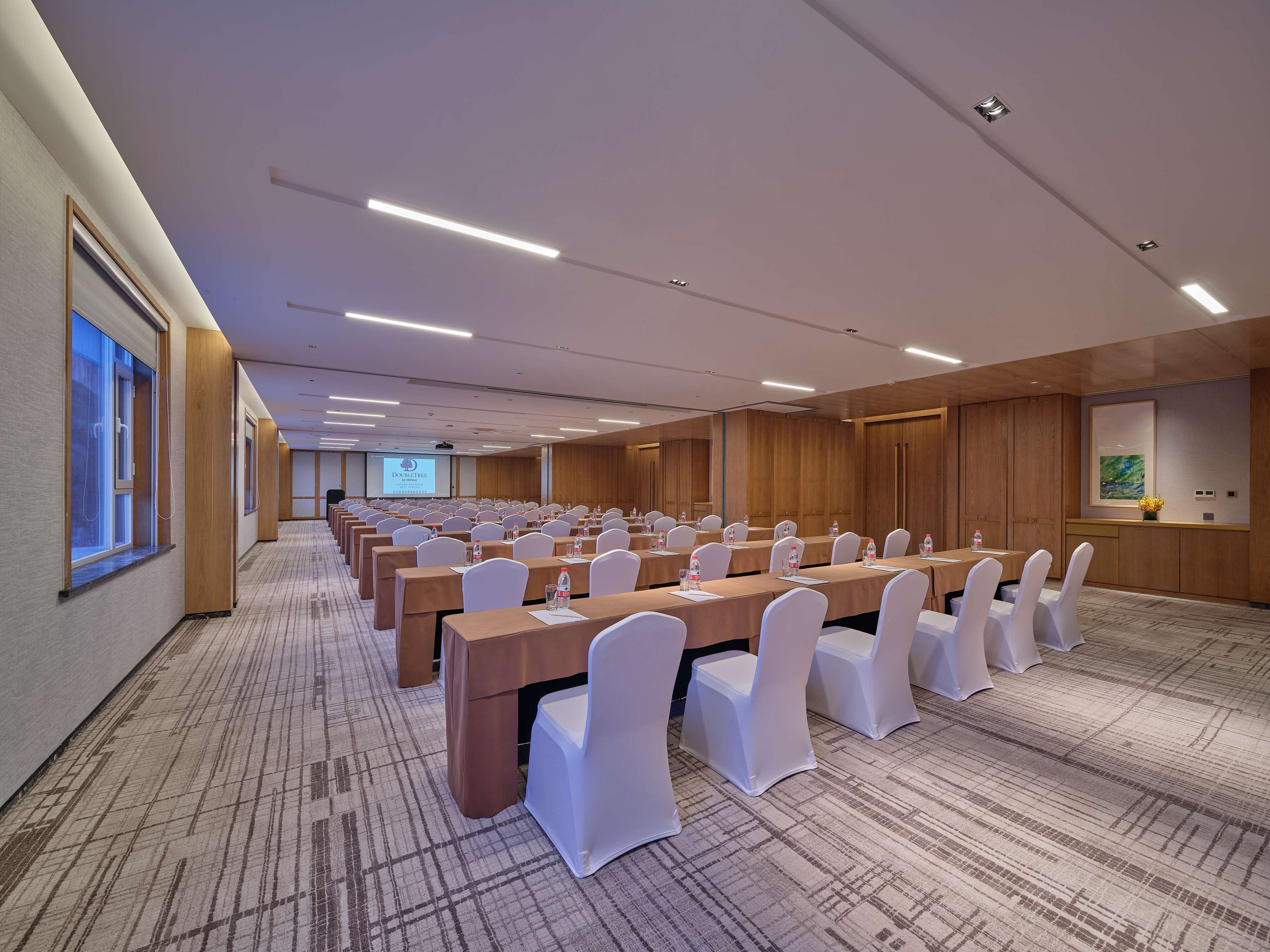 A conference room with rows of tables and chairs, a projector screen, and a wooden panel wall.