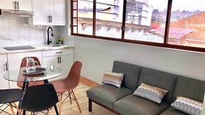 Une cuisine moderne avec des meubles blancs, un robinet noir et une table en verre entourée de chaises. On y trouve également un canapé gris avec des coussins à motifs et une fenêtre donnant sur des immeubles.