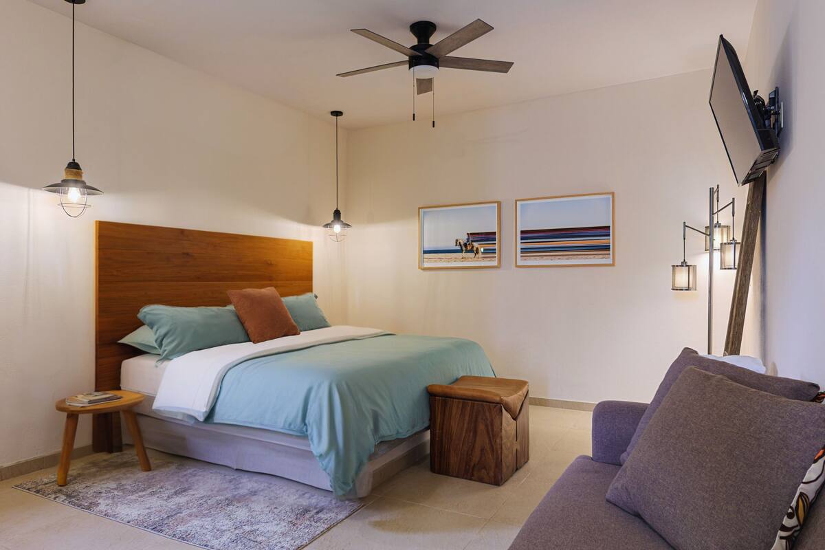 A modern bedroom with a wooden headboard, a bed with teal bedding, a small wooden stool, a purple sofa, and a ceiling fan.
