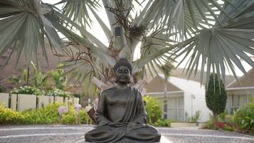 A statue of a seated figure under a palm tree in a landscaped area with a building in the background.