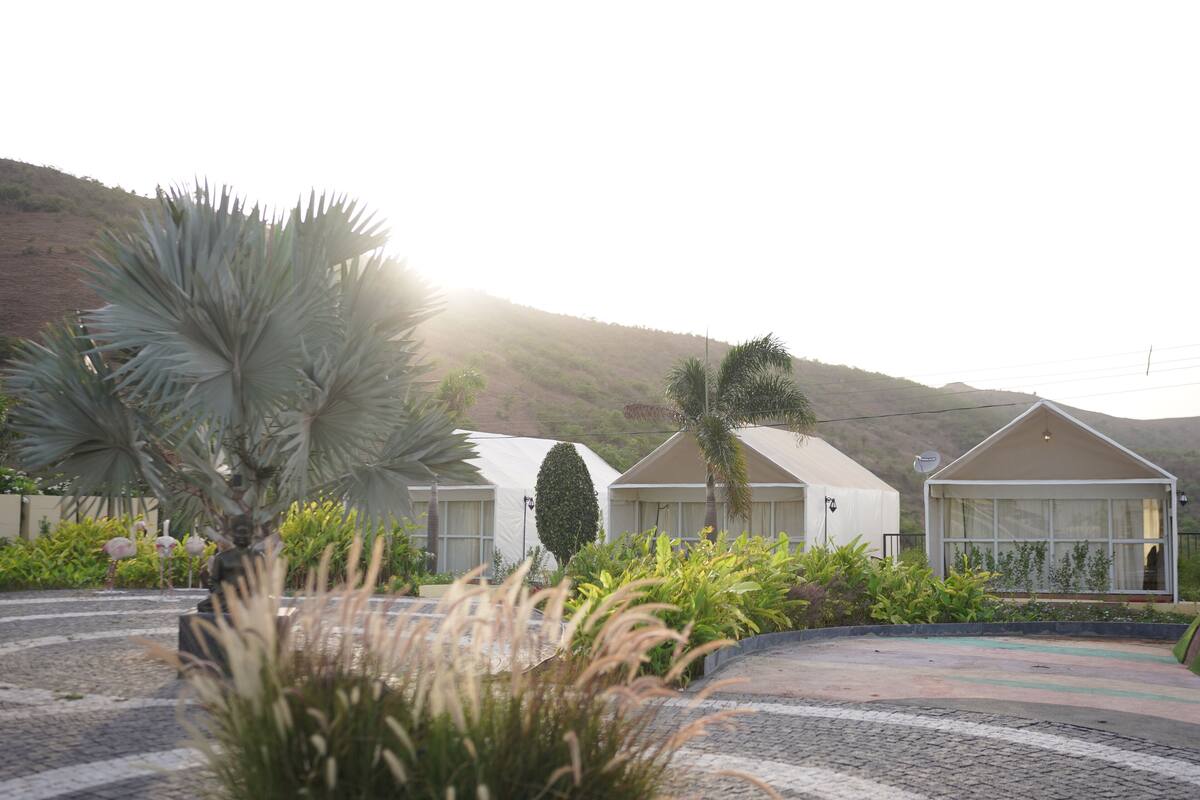A resort with multiple bungalow-style accommodations, surrounded by landscaped gardens and palm trees, set against a hilly backdrop.