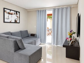 Una sala de estar moderna con un sofá seccional gris, un televisor de pantalla plana y un comedor visible a través de una puerta de vidrio.