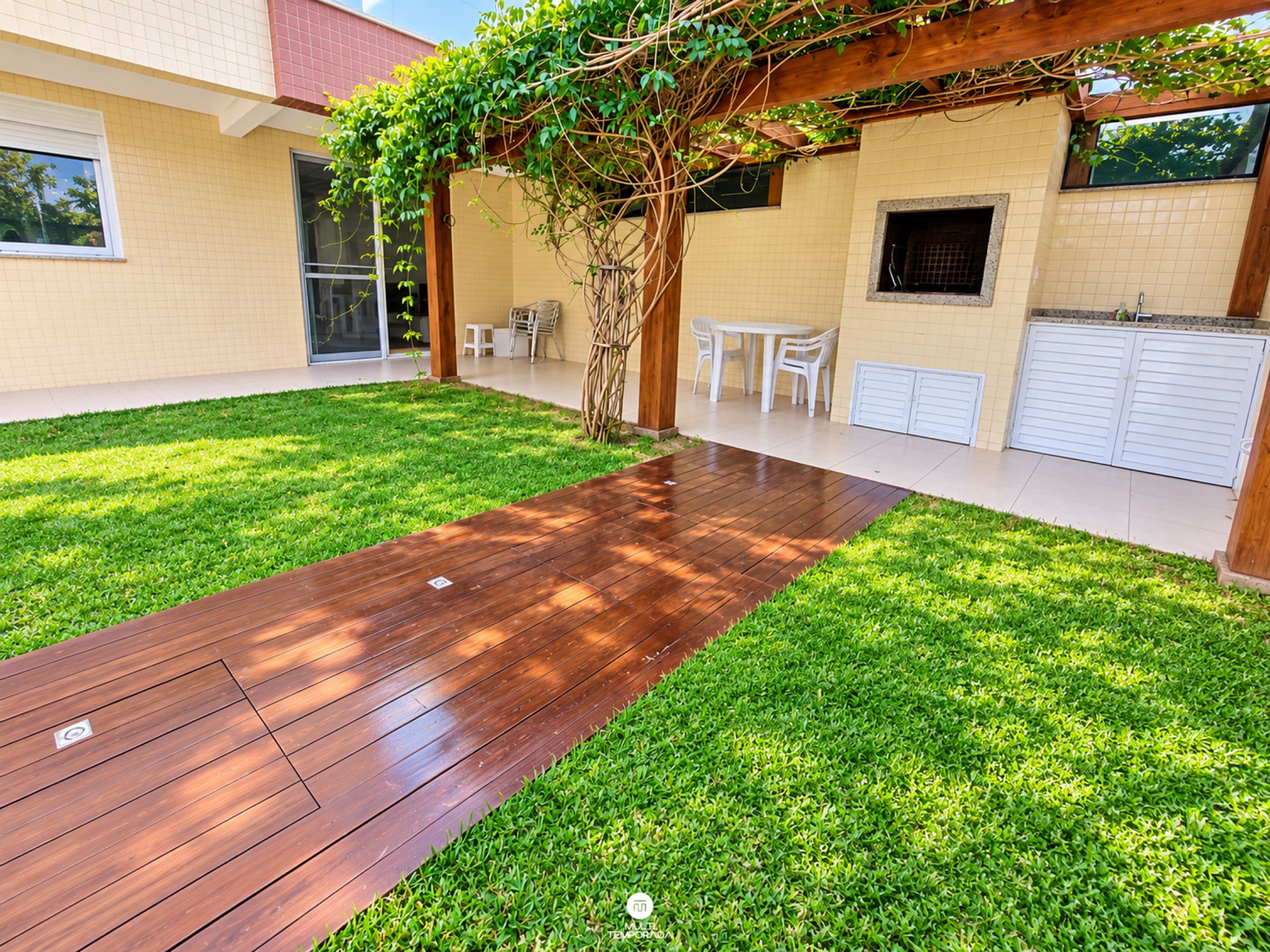 Um quintal com um deck de madeira, gramado, pérgola coberta por plantas trepadeiras, uma mesa e cadeiras brancas, e uma garagem.
