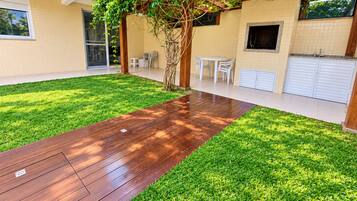 Um quintal com um deck de madeira, gramado, pérgola coberta por plantas trepadeiras, uma mesa e cadeiras brancas, e uma garagem.