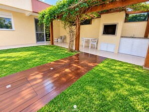 A backyard with a wooden deck, a grassy lawn, a pergola with climbing plants, a white table and chairs, and a garage. - Apartamento com Piscina nos Açores (Florianópolis)