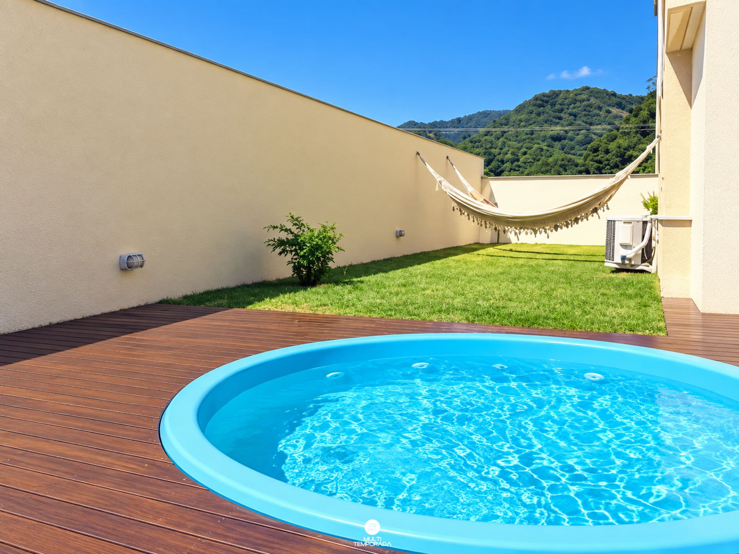 Piscina externa com deck de madeira, rede e área gramada, com montanha ao fundo.