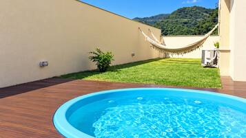 Piscina externa com deck de madeira, rede e área gramada, com montanha ao fundo.