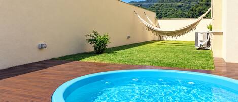 An outdoor swimming pool with a wooden deck, a hammock, and a grassy area with a mountain in the background.