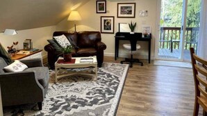 A living room with a brown leather sofa, a coffee table, a desk with a chair, and a large window with a view of greenery. - Happy Valley Studio - Peaceful rest in nature, just 5 minutes from downtown!  (Anacortes)