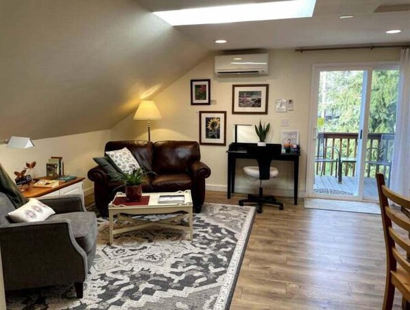 A living room with a brown leather sofa, a coffee table, a desk with a chair, and a large window with a view of greenery.