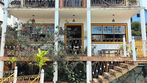 Un edificio de dos pisos con techo de tejas, balcones con barandillas de metal, piscina y plantas en macetas.