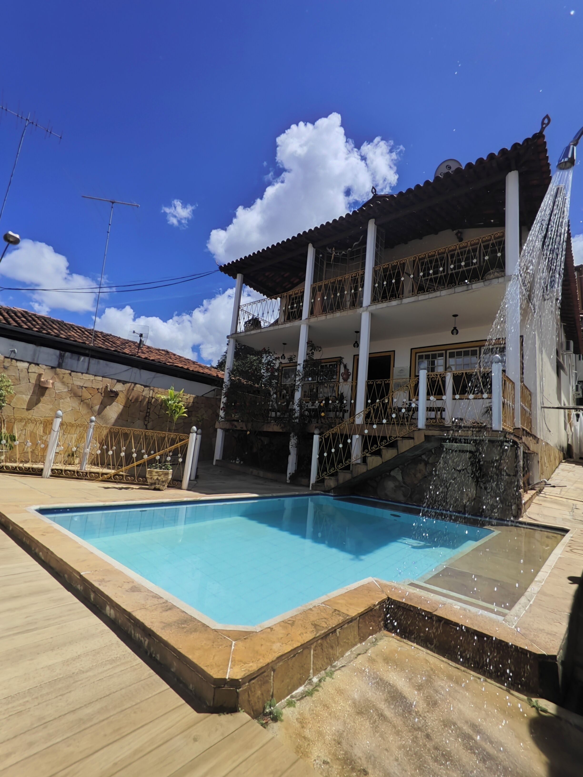 A swimming pool with a wooden deck, a two-story house with a balcony, and a stone wall in the background.