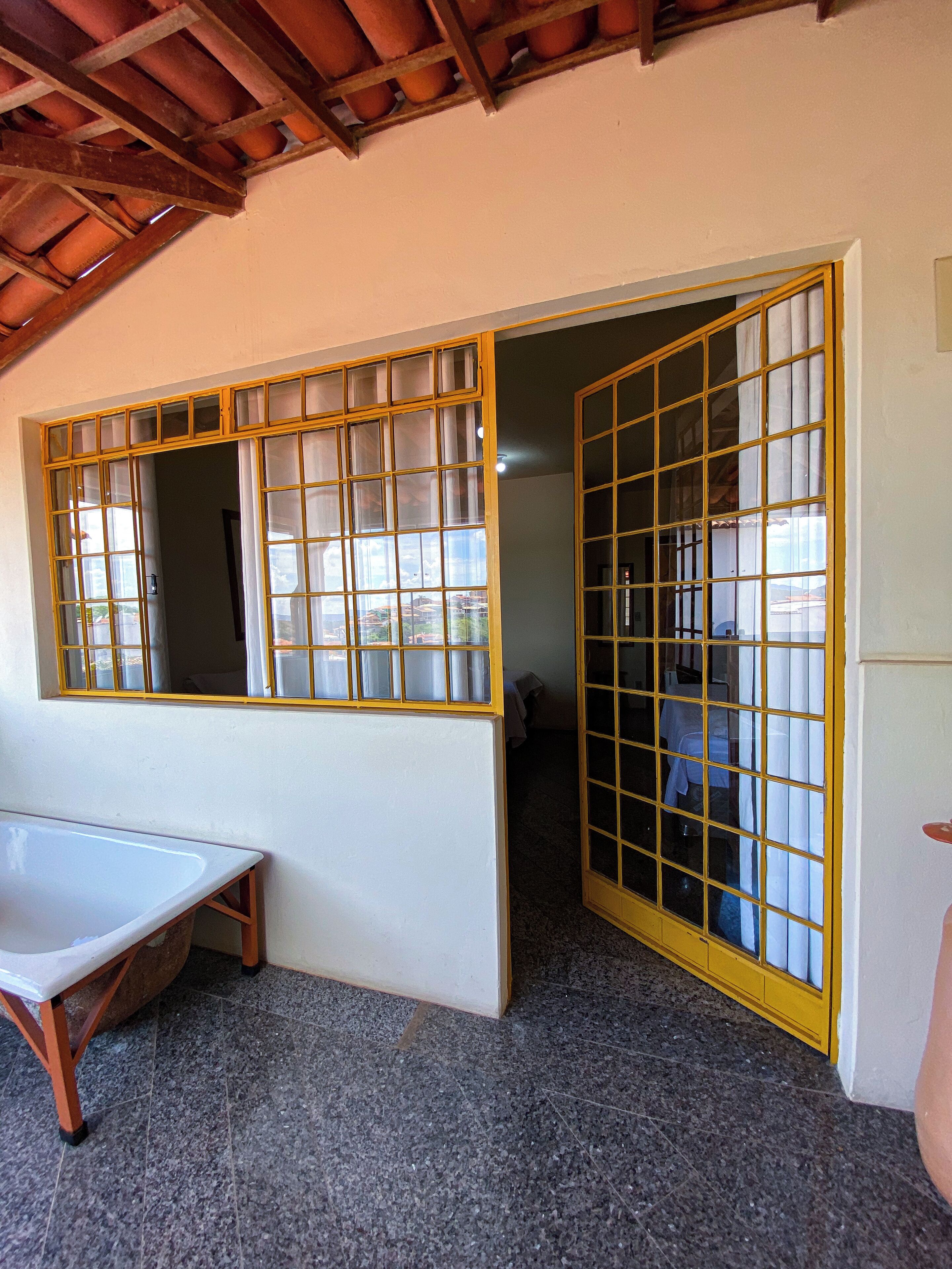 A room with a large window with yellow frames, a white bathtub, and a tiled floor.