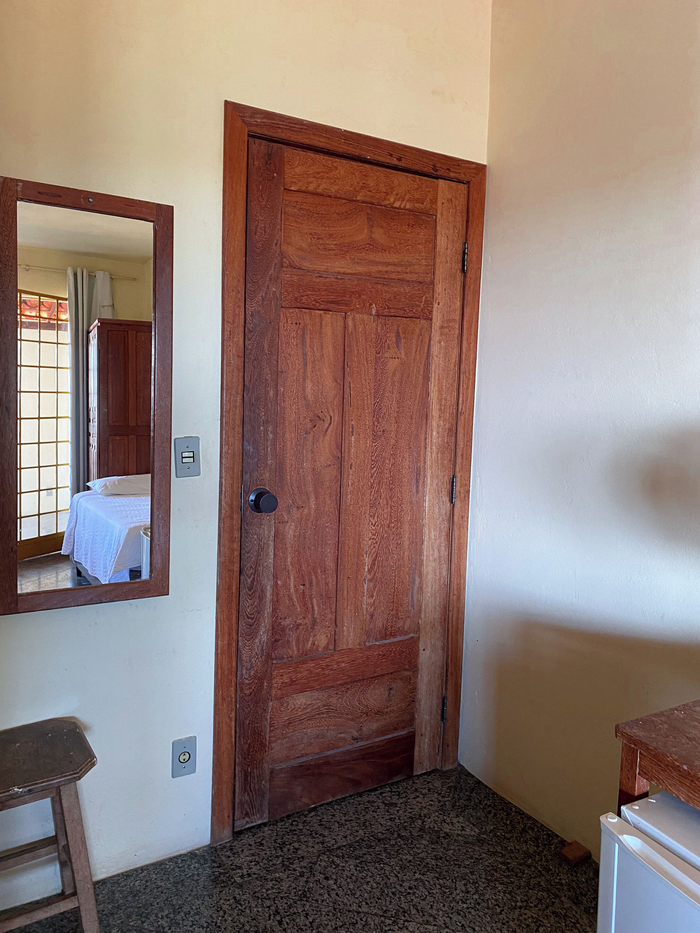 Una puerta de madera con un espejo en la pared, una cama con cubrecama blanco y un pequeño taburete de madera.